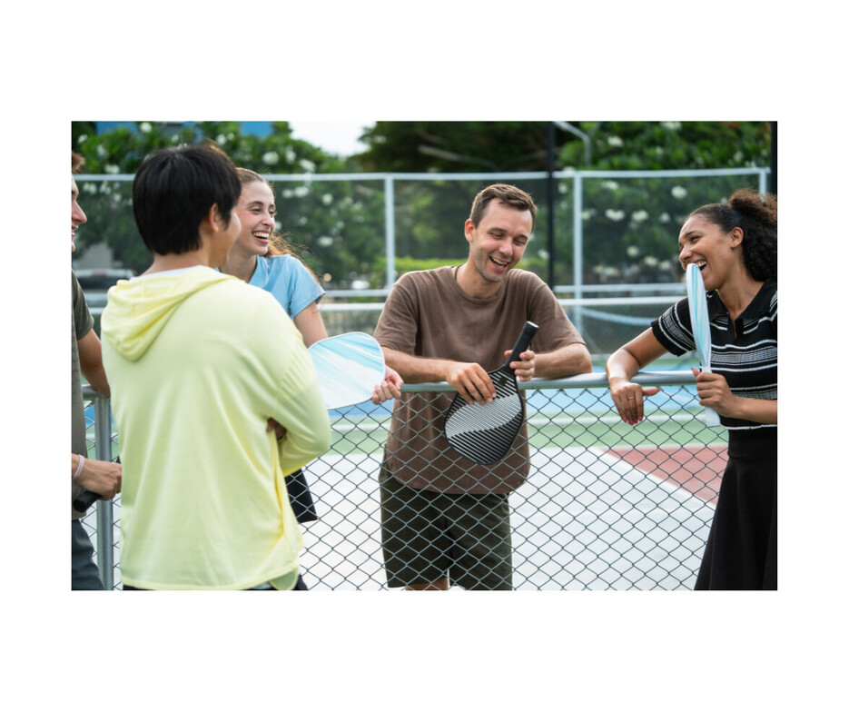 Friends laughing together on an outdoor pickleball court before a game. Staying active and preparing properly can help reduce the risk of pickleball injuries.