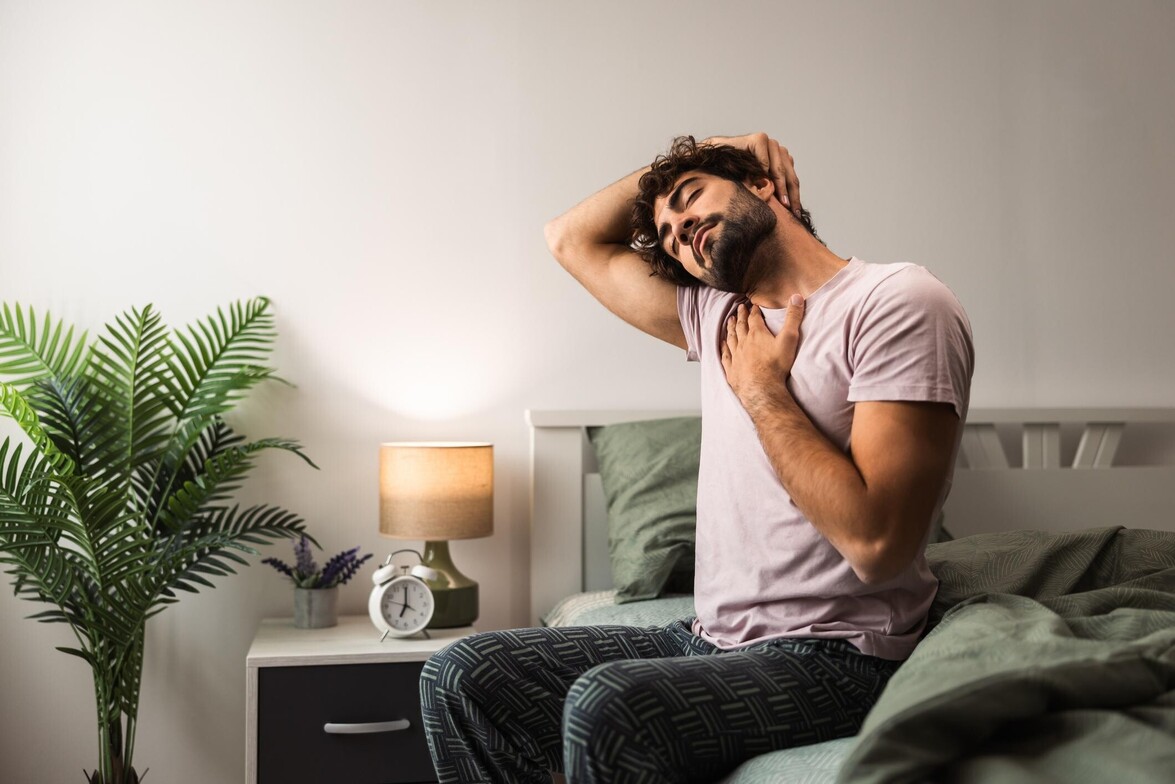 Man sitting on the edge of his bed gently stretching his neck to relieve morning stiffness.